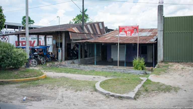 Weathered low roadside shops with corrugated roofs, hand-painted signs, open bays, and a parked motorcycle.