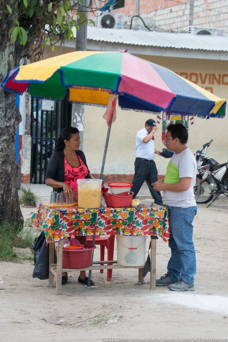 Woman serving at a roadside food stand under a multicolored umbrella with containers on a floral tablecloth.