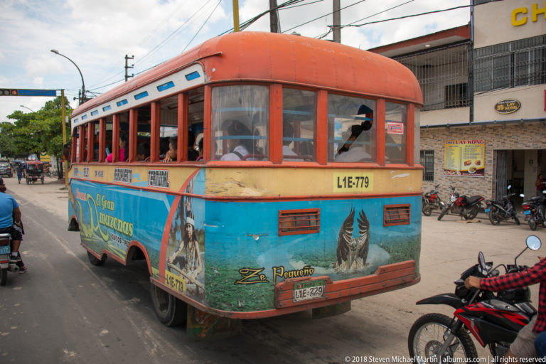Rear view of orange-and-turquoise local bus with open windows traveling among motorcycles in Iquitos.
