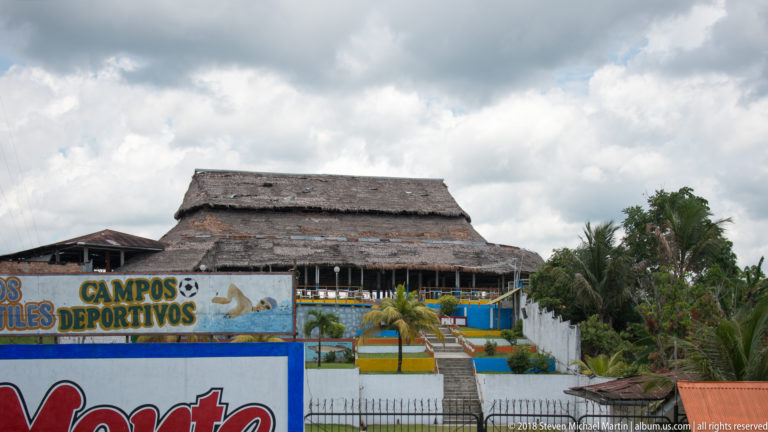 Large thatched-roof recreational structure behind signs referring to sports fields, with palms and fenced grounds.