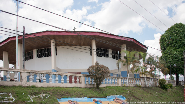 Large open-sided building with white balustrade, regular columns, and a broad red-edged roof.