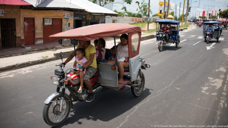 Woman driving a red-canopied mototaxi carrying several children along a city street.