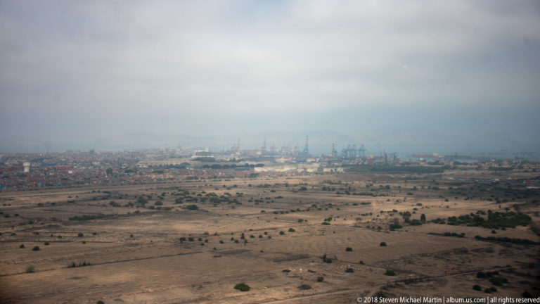 Aerial view of an arid coastal plain with distant city development and port cranes in haze.