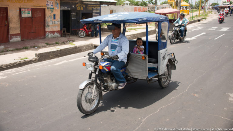 Blue-canopied mototaxi approaching with an adult driver and small child seated in the rear.
