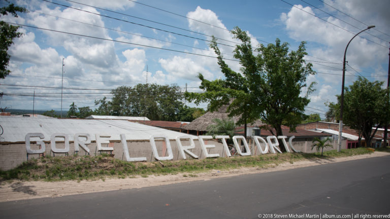 Large white letters spelling “Loreto” beside a paved Iquitos road under a bright cloudy sky.