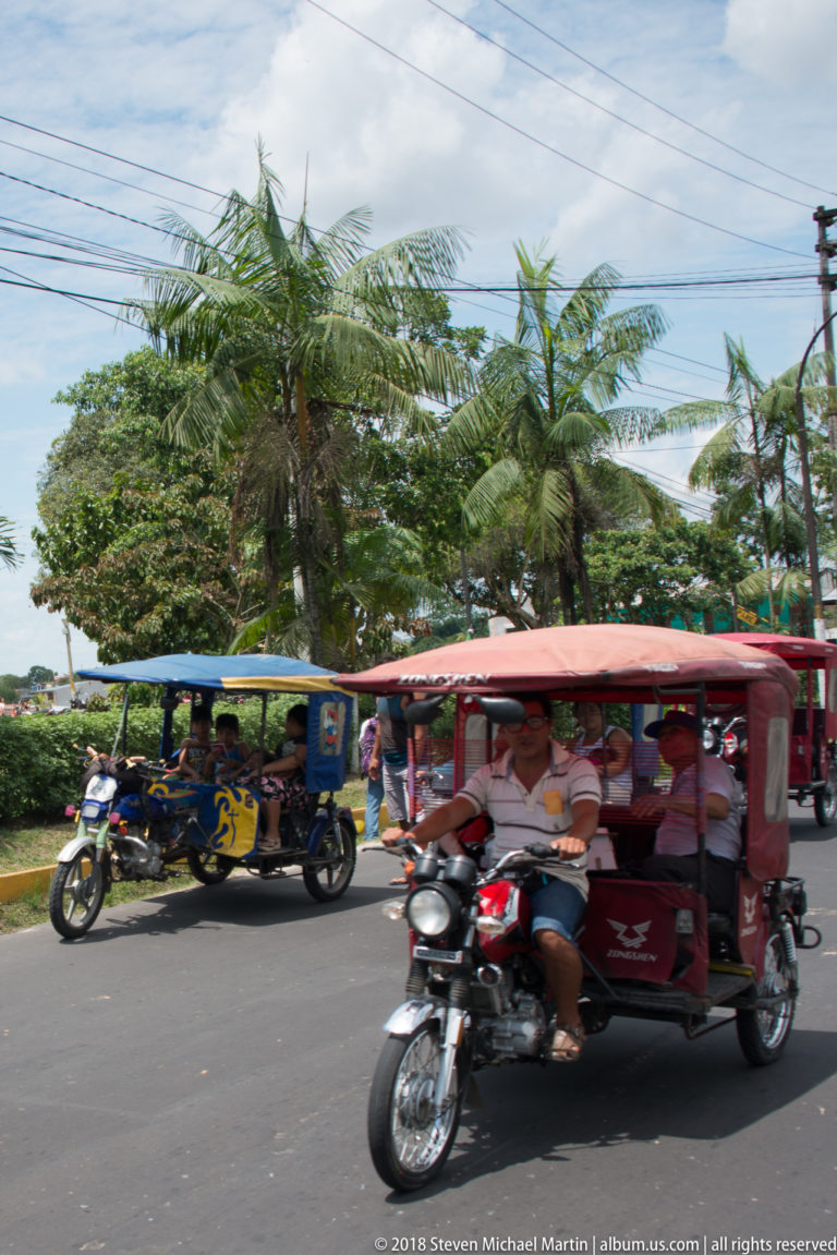 Red mototaxi moving past a landscaped roadside area with palms and other mototaxis behind.