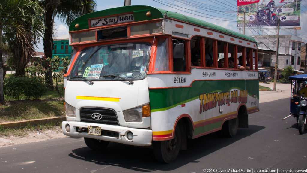 White, green, and orange local bus with open side windows traveling along an Iquitos street.