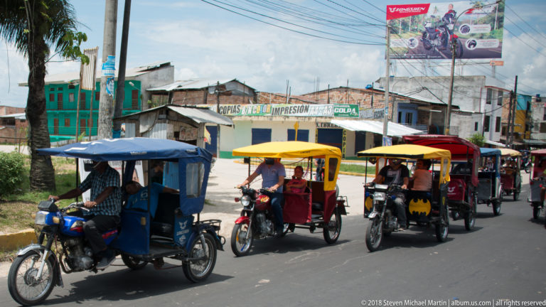 Blue, yellow, red, and dark-canopied mototaxis traveling in a line along an Iquitos street.