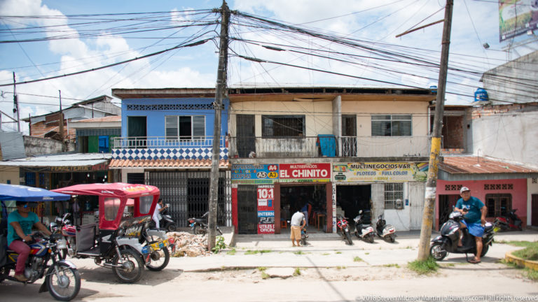 Two-story neighborhood shops with signs, motorcycles, mototaxis, and dense overhead utility wires.