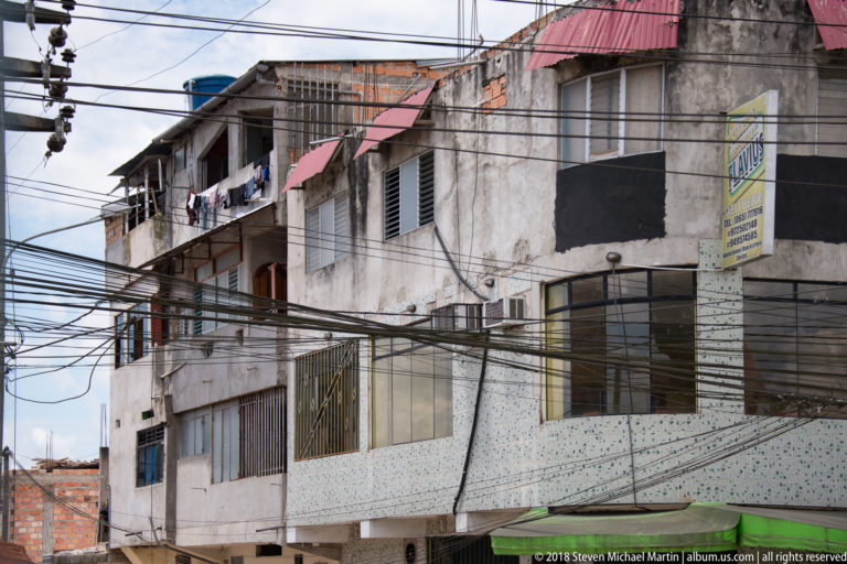 Weathered multi-story residential block with barred windows, hanging laundry, roof shades, and dense overhead wiring.