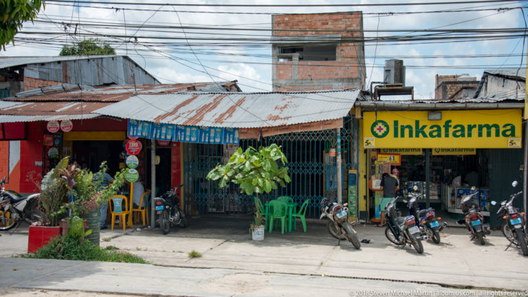 Inkafarma pharmacy beside low neighborhood shops with motorcycles parked across the forecourt.