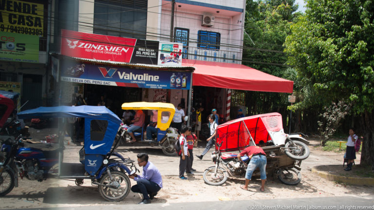 Mototaxis outside a lubricant shop, including one red three-wheeler tilted up for roadside repair.