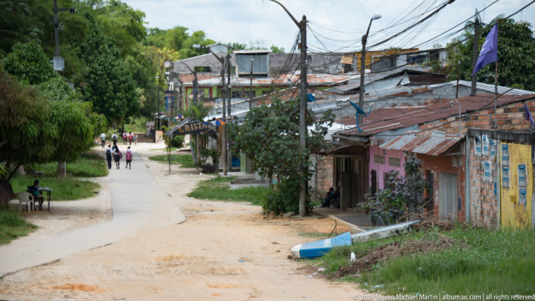 Quiet neighborhood road with modest houses, sandy verges, corrugated roofs, and dense overhead utility wires.
