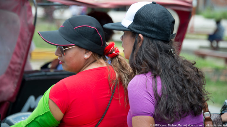 Two women in profile beside the open side of a red mototaxi, one wearing a black cap.