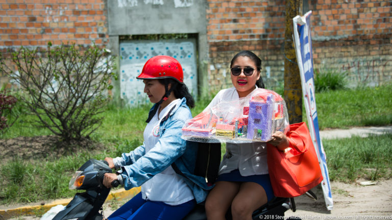 Woman carrying a tray of small packaged goods while riding as a passenger on a motorcycle.