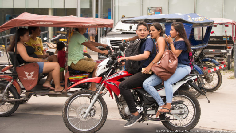 Three young women riding together on a red motorcycle amid mototaxis and other street traffic.
