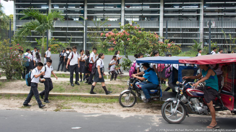 Uniformed students walking outside Colegio Nacional Iquitos beside blue and red mototaxis.