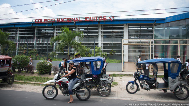 Students and mototaxis outside the fenced frontage of Colegio Nacional Iquitos beneath palm trees.