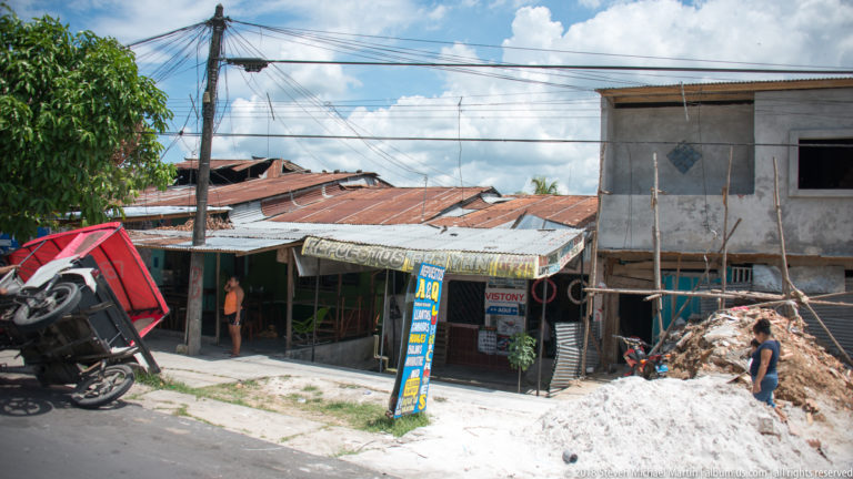 Large sand pile outside corrugated-roof shops with motorcycles, a mototaxi, signs, and a worker nearby.