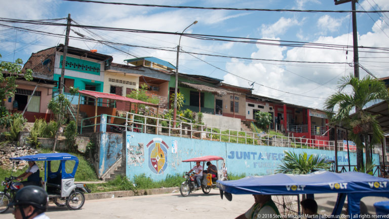 Colorful hillside homes above a painted wall with palms, utility wires, and mototaxis along the street.