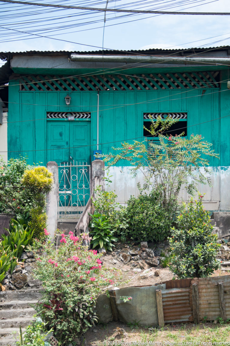 Turquoise wooden house behind flowering shrubs, with shuttered openings and a small raised entrance.