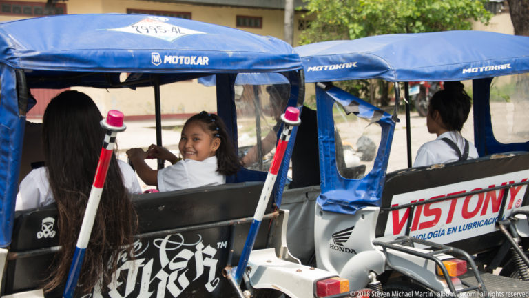 School-age girls riding in the open rear of a blue-canopied mototaxi, one smiling toward the camera.