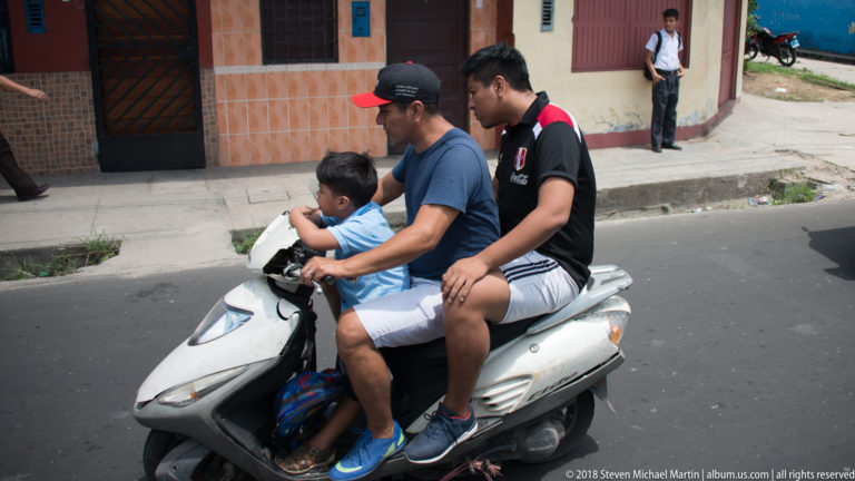 Adult driving a white scooter with a child seated in front and a second passenger riding behind.