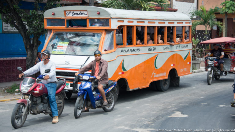 Orange city bus with open windows traveling beside two motorcycles and a mototaxi.