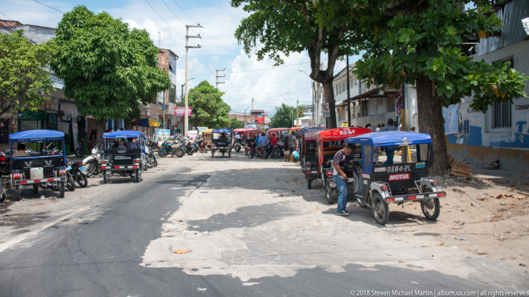 Mototaxis lining both sides of a shaded Iquitos street beneath trees and vendor umbrellas.
