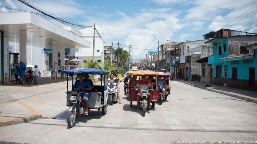 Two mototaxis approaching on a sunlit street with motorcycles, a service station, and low-rise buildings.