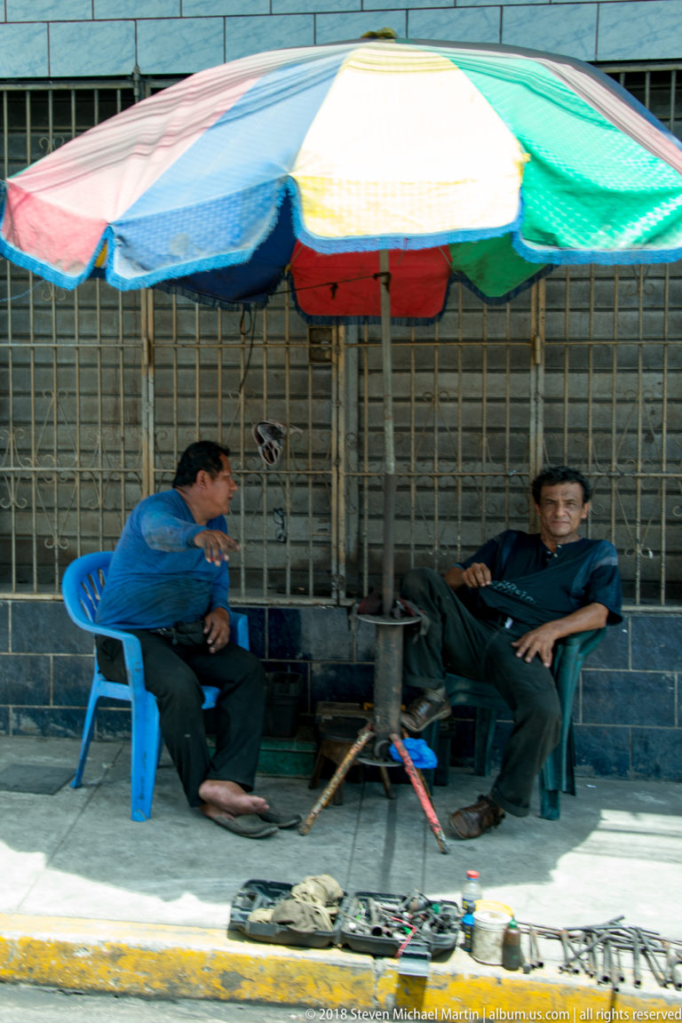 Two men sitting beneath a multicolored umbrella beside small goods displayed on the sidewalk.