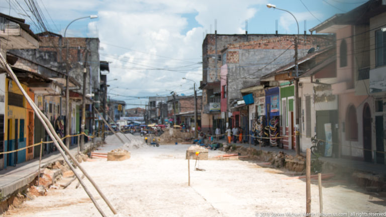 Street under reconstruction with pale roadbed, trenches, stakes, debris, and low-rise buildings on both sides.
