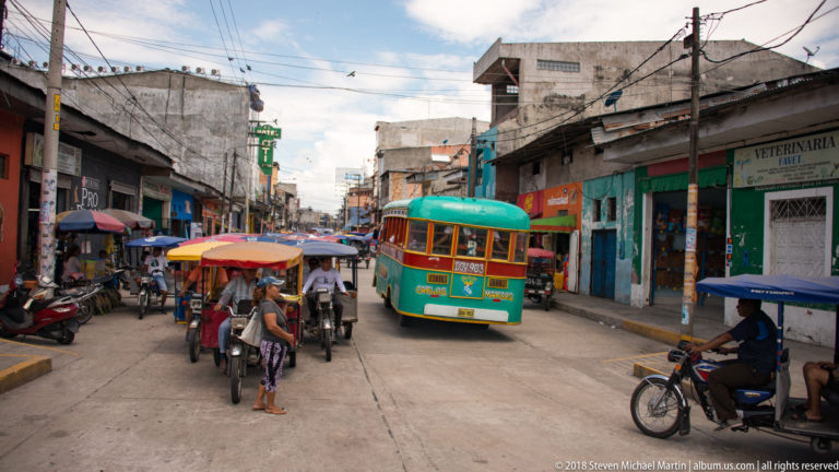 Green-and-orange bus moving through a busy Iquitos street among mototaxis, motorcycles, and pedestrians.