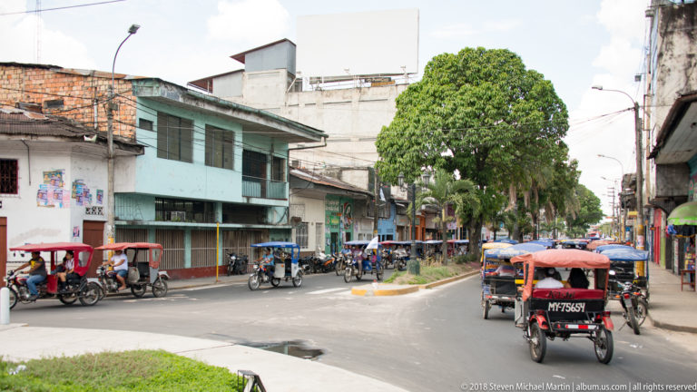 Mototaxis traveling along a divided Iquitos street with low-rise buildings and a planted median.