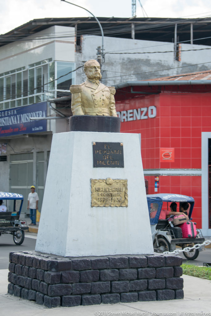 Bust on a pale stone pedestal with plaques, set beside storefronts and passing mototaxis.