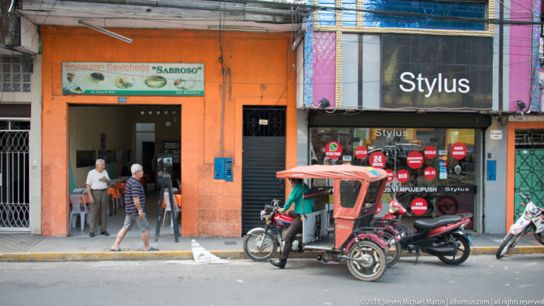 Orange cevichería beside a dark Stylus storefront, with a red mototaxi and motorcycles at the curb.