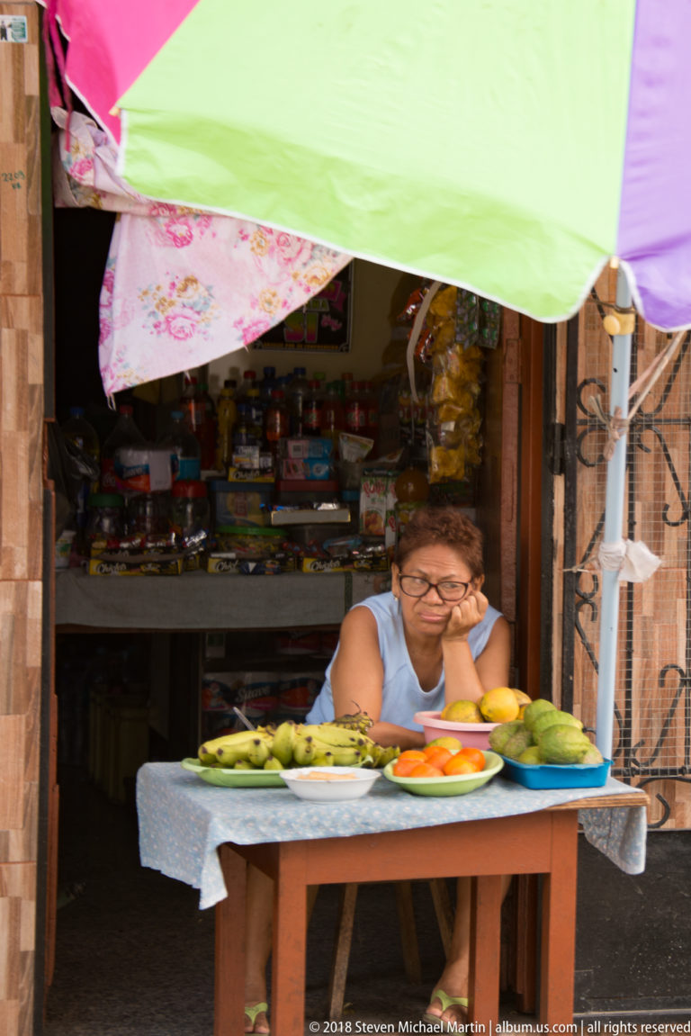 Woman seated behind fruit and produce at a shop doorway shaded by overlapping umbrellas.