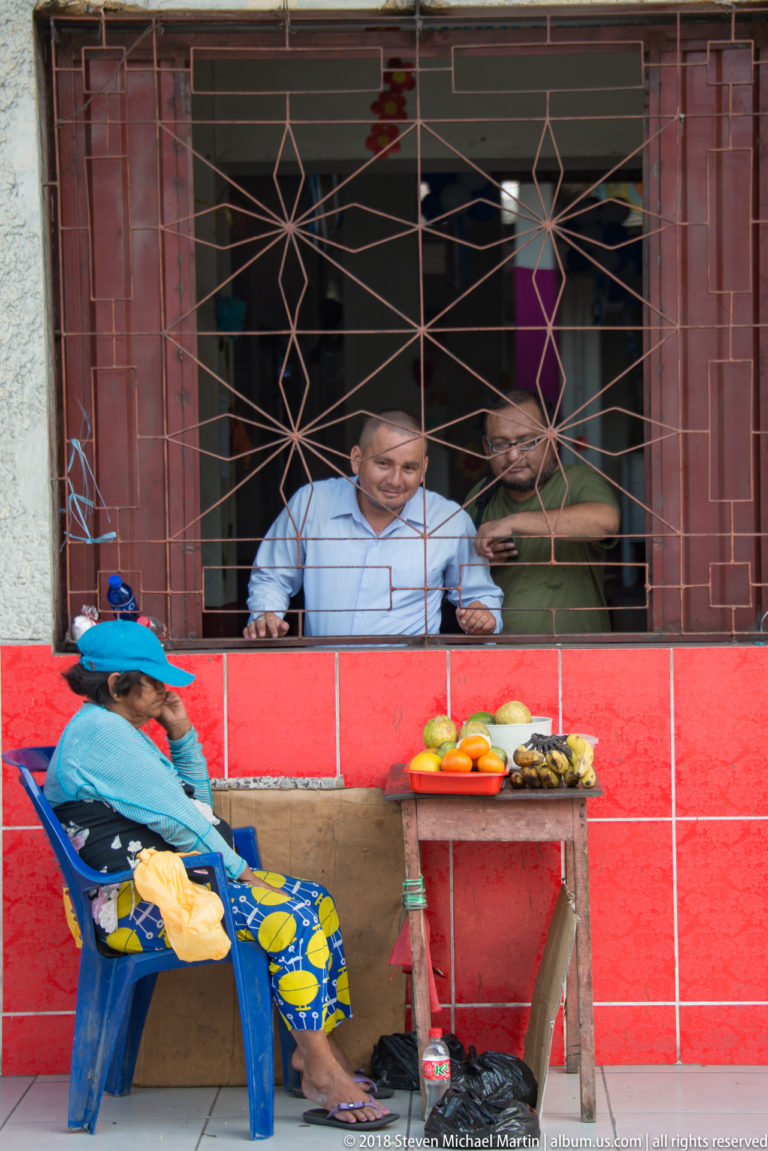 Seated woman beside a small fruit table under a barred window with two men standing behind it.