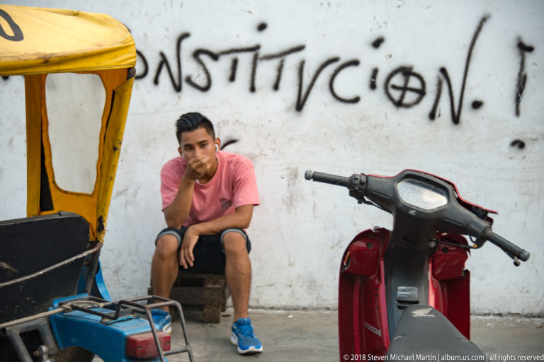 Young man seated against a graffiti-marked wall between a red scooter and part of a mototaxi.