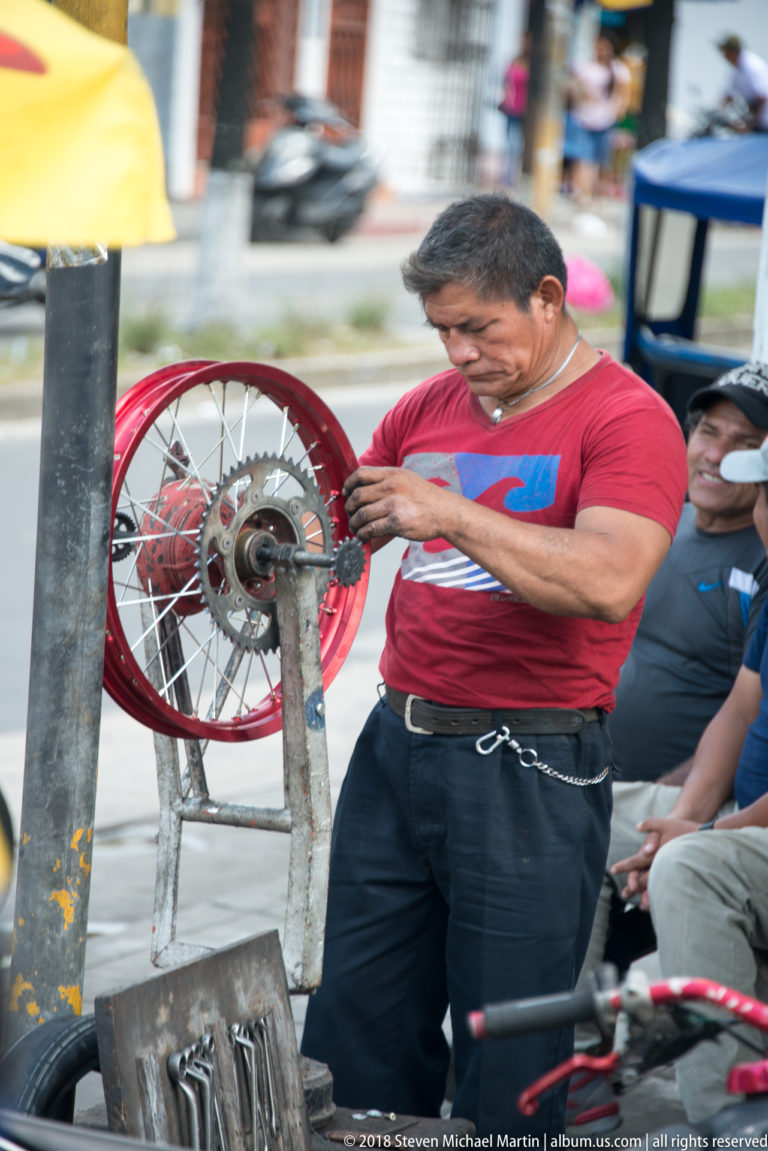 Man repairing the hub of a red-rimmed motorcycle wheel at a curbside work station.