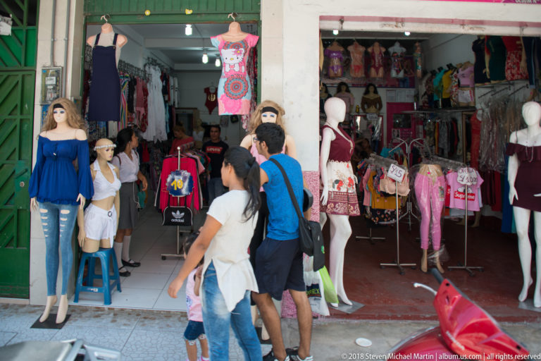 Open clothing shop with mannequins in dresses, racks of garments, and shoppers passing the entrance.