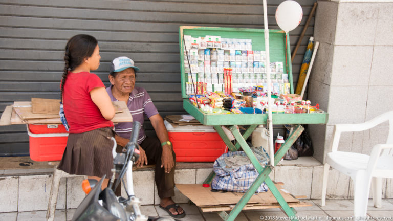 Two women beside a small green sidewalk display filled with packaged goods and accessories.