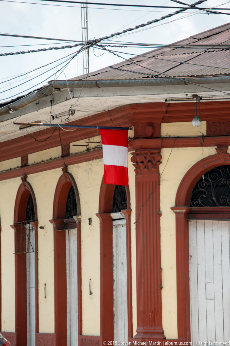 Peruvian flag hanging from a cream-and-red façade with arched openings and iron transoms.