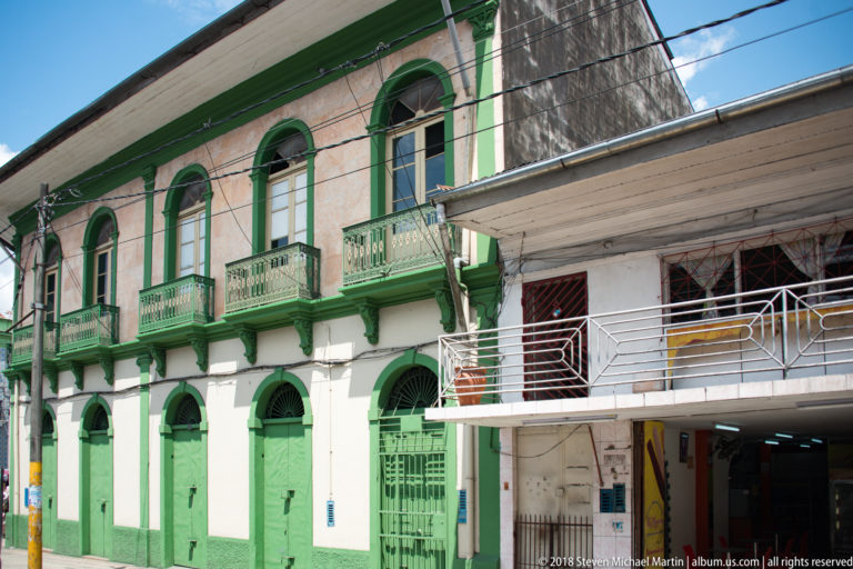 Green-and-white two-story building with arched doors, narrow balconies, and overhead utility wires.