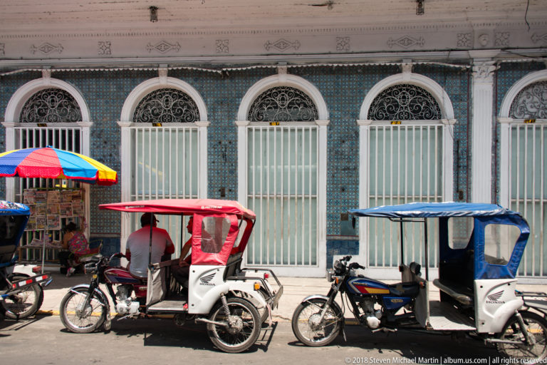 Blue tiled façade with repeated arched doors and iron transoms, partly screened by mototaxis and a street stall.