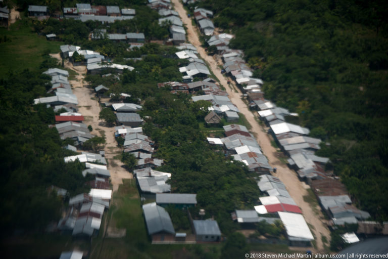 Aerial view of houses lining a straight unpaved road through dense tropical forest.