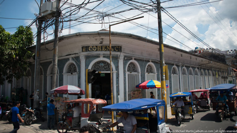 Long Cohen storefront with arched ironwork windows, mototaxis, and umbrella-covered vendors along the curb.