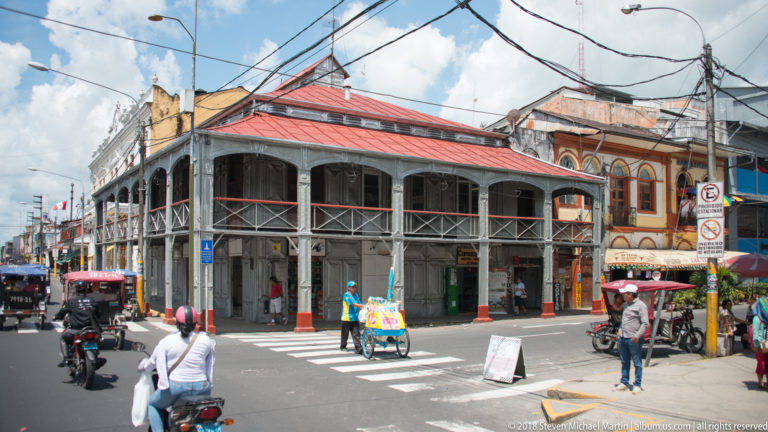 Busy Iquitos intersection with a two-story metal arcaded building, mototaxis, pedestrians, and overhead wires.