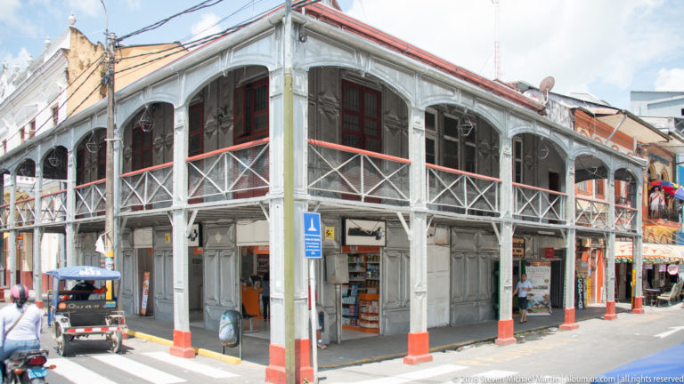 Two-story corner building with metal columns, arched openings, cross-braced galleries, and street-level shops.