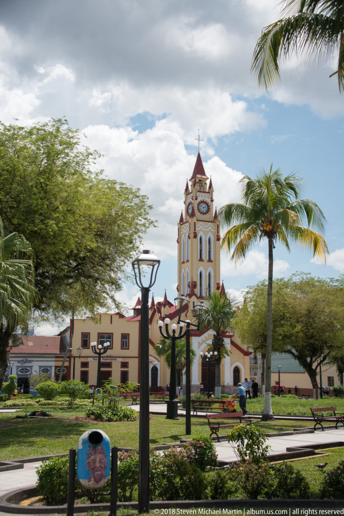 Landscaped Plaza de Armas with palms and lamps facing Iquitos Cathedral and its clock tower.
